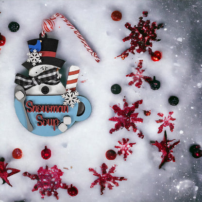 Festive scene featuring Janet's Craft Corner Snowman Soup Ornament, a DIY craft kit shaped like a mug with a snowman and candy cane handle. Red and black snowflakes adorn the snowy backdrop, complemented by red berries. Perfect for holiday décor, it includes a charming bow tie detail.