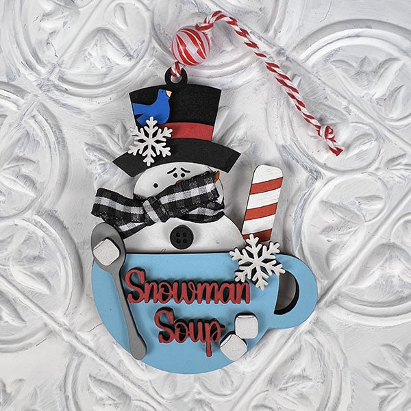 The Snowman Soup Ornament from Janet's Craft Corner, an ideal choice for home décor, features a snowman inside a blue mug topped with a black hat and red band, adorned with snowflakes. A candy cane and striped scarf enhance its charm, while a twisted red and white string allows it to hang elegantly against any patterned backdrop.