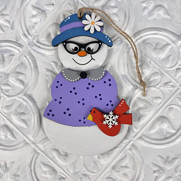 This delightful decoration from Janet's Craft Corner’s Vintage Snow Couple Ornaments collection showcases a snowman adorned in a purple dress, a blue hat with a white flower, and glasses. It is holding a red bird with a snowflake against its textured white backdrop. Perfect for any hand-painted ornaments collection, it includes a convenient string for hanging.