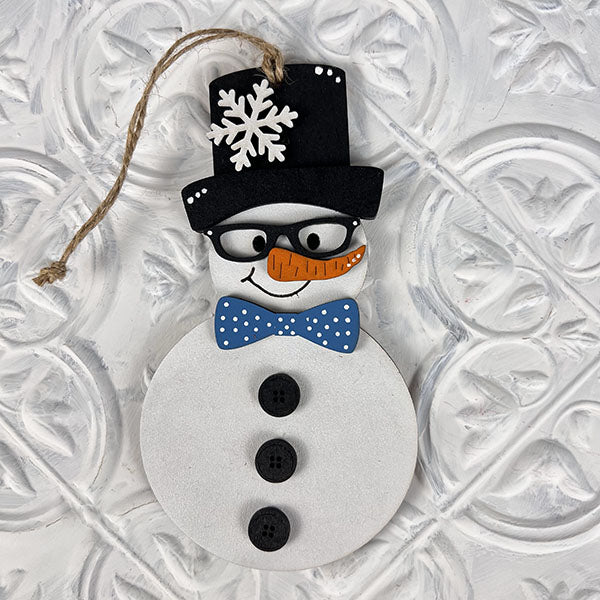 Experience the charm of Janet's Craft Corner with the Vintage Snow Couple Ornaments - Painted. This delightful hand-painted snowman comes adorned with a top hat, glasses, a carrot nose, and an eye-catching blue polka dot bow tie. It features three black buttons and includes a looped string for easy hanging against any textured white surface.