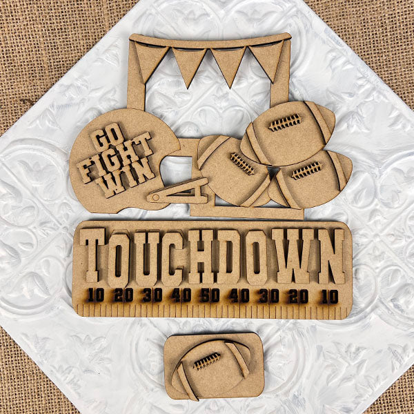 Transform your space with Janet's Craft Corner's Football Interchangeable Insert DIY home decor kit. This hand-painted set features wooden cutouts, including a football helmet inscribed with "Go Fight Win," four footballs, and a “Touchdown” sign above a yard line. It also comes with an additional smaller football insert at the bottom – perfect for adding a personalized touch to your decor.