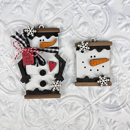 Two Marshmallow Snowman Ornaments from Janet's Craft Corner's DIY Decoration Kits are set against a textured white background. One ornament is decorated with a black and white checkered ribbon and a red tag, while the other showcases a carrot nose and snowflake details. Both ornaments feature buttons and wooden accents, making them an ideal addition to any holiday decor collection.