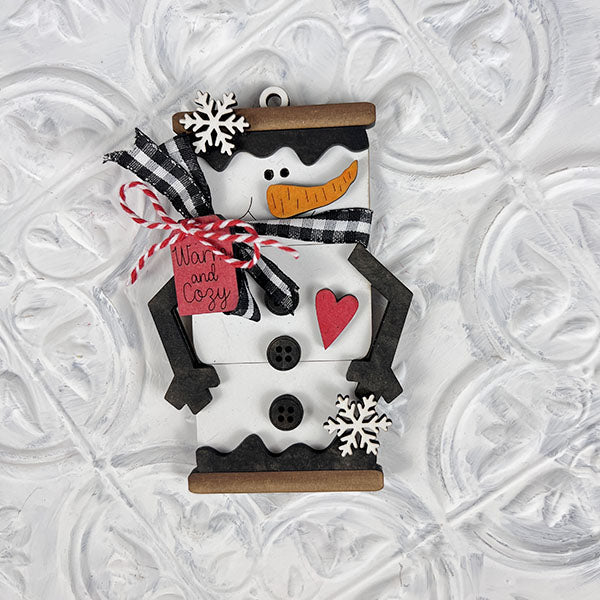 Introducing the delightful Marshmallow Snowman Ornaments from Janet's Craft Corner! These DIY decoration kits feature adorable snowmen with carrot noses, black button eyes, and a charming red heart on their bellies. Each snowman is adorned with a stylish black and white plaid scarf, a "Warm and Cozy" tag, and playful snowflake accents, making them perfect for your holiday decor against a textured white backdrop.