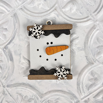 The Marshmallow Snowman Ornaments from Janet's Craft Corner, perfect for holiday decor, showcase a melting snowman's face complete with a carrot nose, two snowflakes, and small black dots for eyes on a white patterned background.