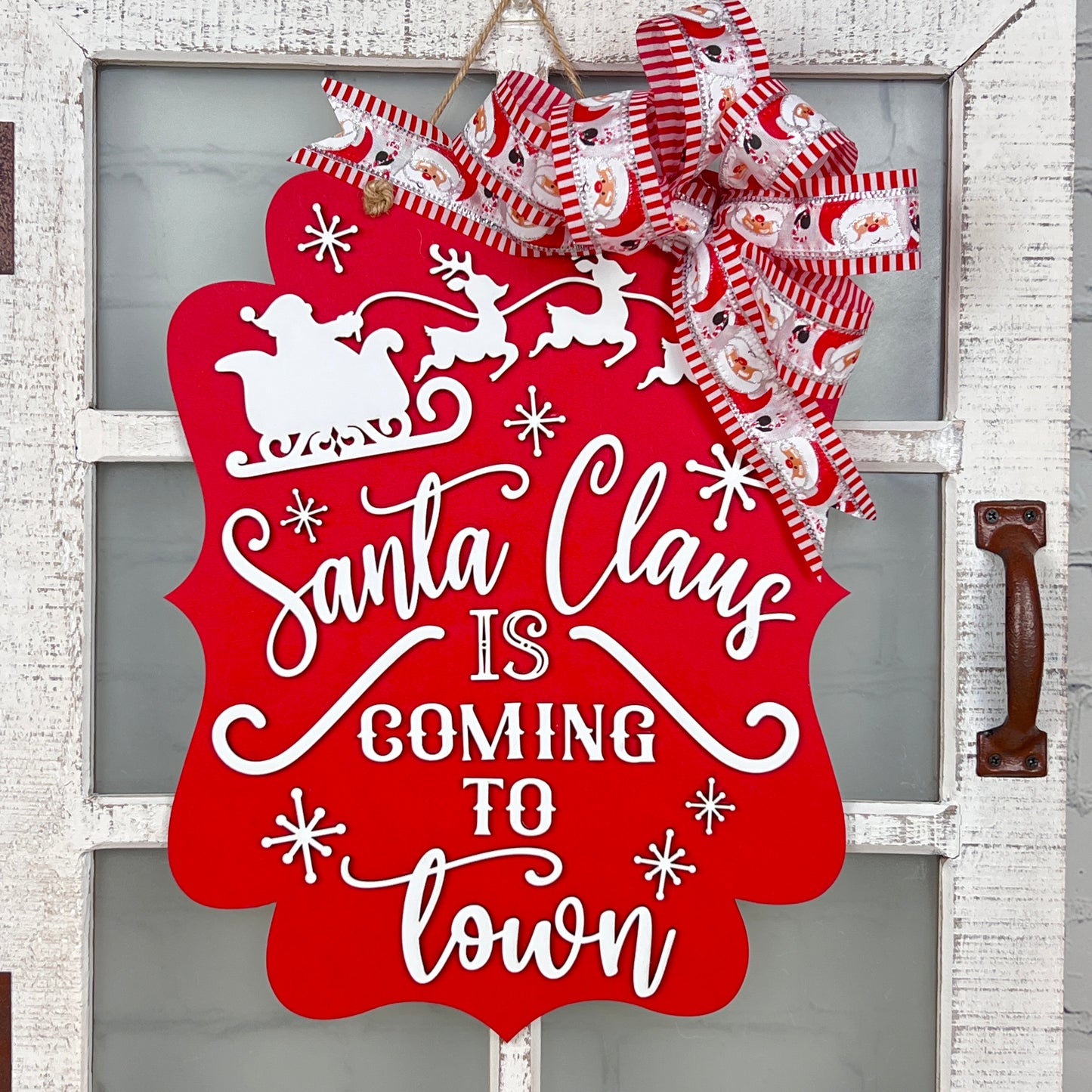 The "Santa is Coming to Town Door Hanger" by Janet's Craft Corner displays a red sign with white lettering, showcasing silhouettes of Santa's sleigh and reindeer. The DIY craft kit comes with a festive red and white ribbon adorned with holiday patterns, making it an ideal addition to any white door.