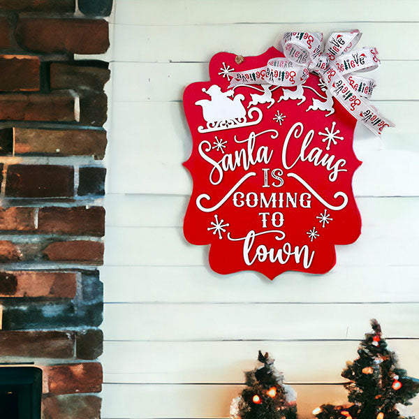 A festive door decoration from Janet's Craft Corner adorns a white wall, featuring the "Santa is Coming to Town Door Hanger" with a red sign depicting Santa's sleigh and reindeer. The sign reads "Santa Claus is Coming to Town" and includes an attached bow with the word "believe." Below, two small decorated Christmas trees by a brick fireplace capture the holiday spirit.