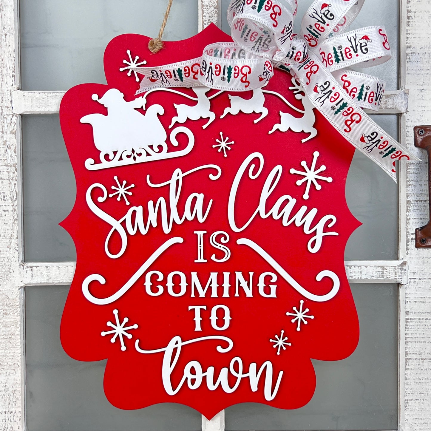 A red "Santa is Coming to Town Door Hanger" from Janet's Craft Corner is displayed on the door, perfectly encapsulating the holiday spirit. The white text features "Santa Claus is Coming to Town," accompanied by a silhouette of Santa in a sleigh at the top, and it's completed with a cheerful bow for a festive touch.