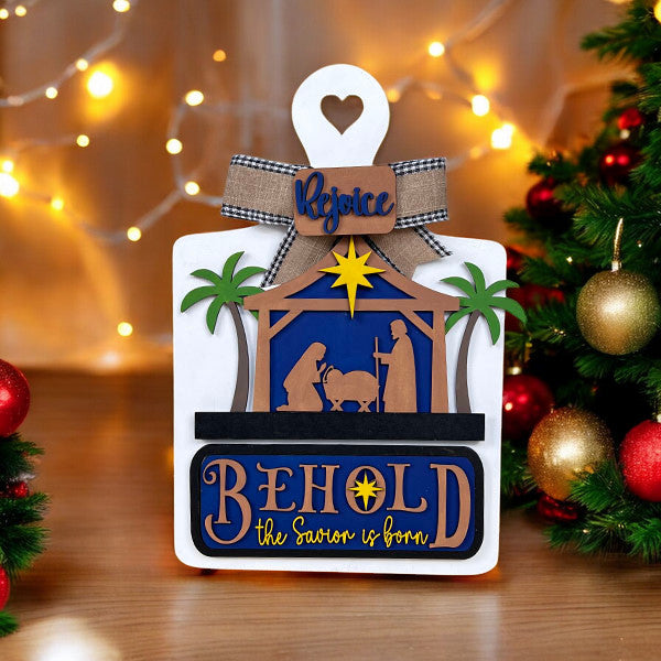 Celebrate the holiday spirit with Janet's Craft Corner's Nativity Interchangeable Insert, featuring Mary, Joseph, and baby Jesus under a star. Decorated with palm trees, Christmas lights, and the phrases "Behold the Savior is born" and "Rejoice," it's a festive addition to any decor.