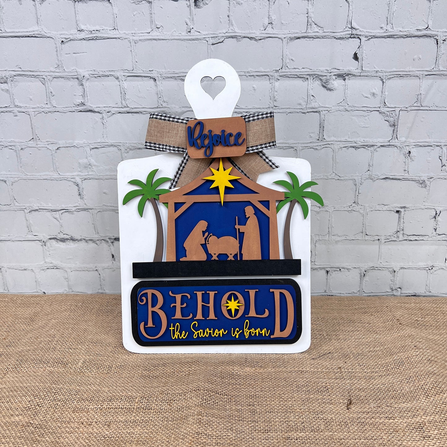 The Nativity Interchangeable Insert from Janet's Craft Corner, a DIY home decor kit, showcases a hand-painted wooden nativity scene adorned with palm trees and the word "Rejoice" at the top. Below, the bold phrase "BEHOLD the Savior is born" highlights Mary, Joseph, and baby Jesus on a burlap surface set against a brick wall background. Ideal for fans of interchangeable home decorations.