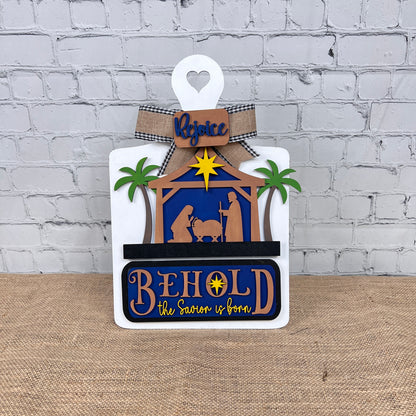 The Nativity Interchangeable Insert from Janet's Craft Corner, a DIY home decor kit, showcases a hand-painted wooden nativity scene adorned with palm trees and the word "Rejoice" at the top. Below, the bold phrase "BEHOLD the Savior is born" highlights Mary, Joseph, and baby Jesus on a burlap surface set against a brick wall background. Ideal for fans of interchangeable home decorations.