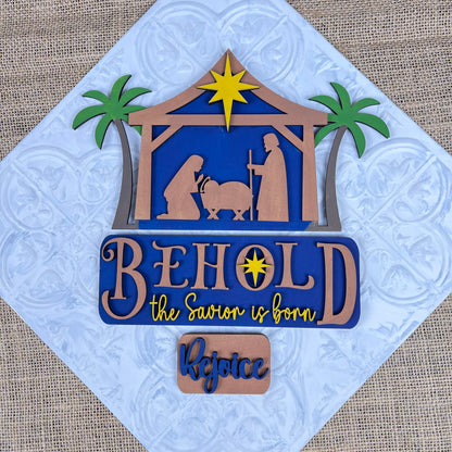 The Nativity Interchangeable Insert from Janet's Craft Corner is an exquisite DIY home decor kit, showcasing a hand-painted scene of Mary, Joseph, and baby Jesus in a manger under a radiant star with palm trees. The piece features the inspiring phrases "Behold, the Savior is born" and "Rejoice," all set against a beautifully textured background.