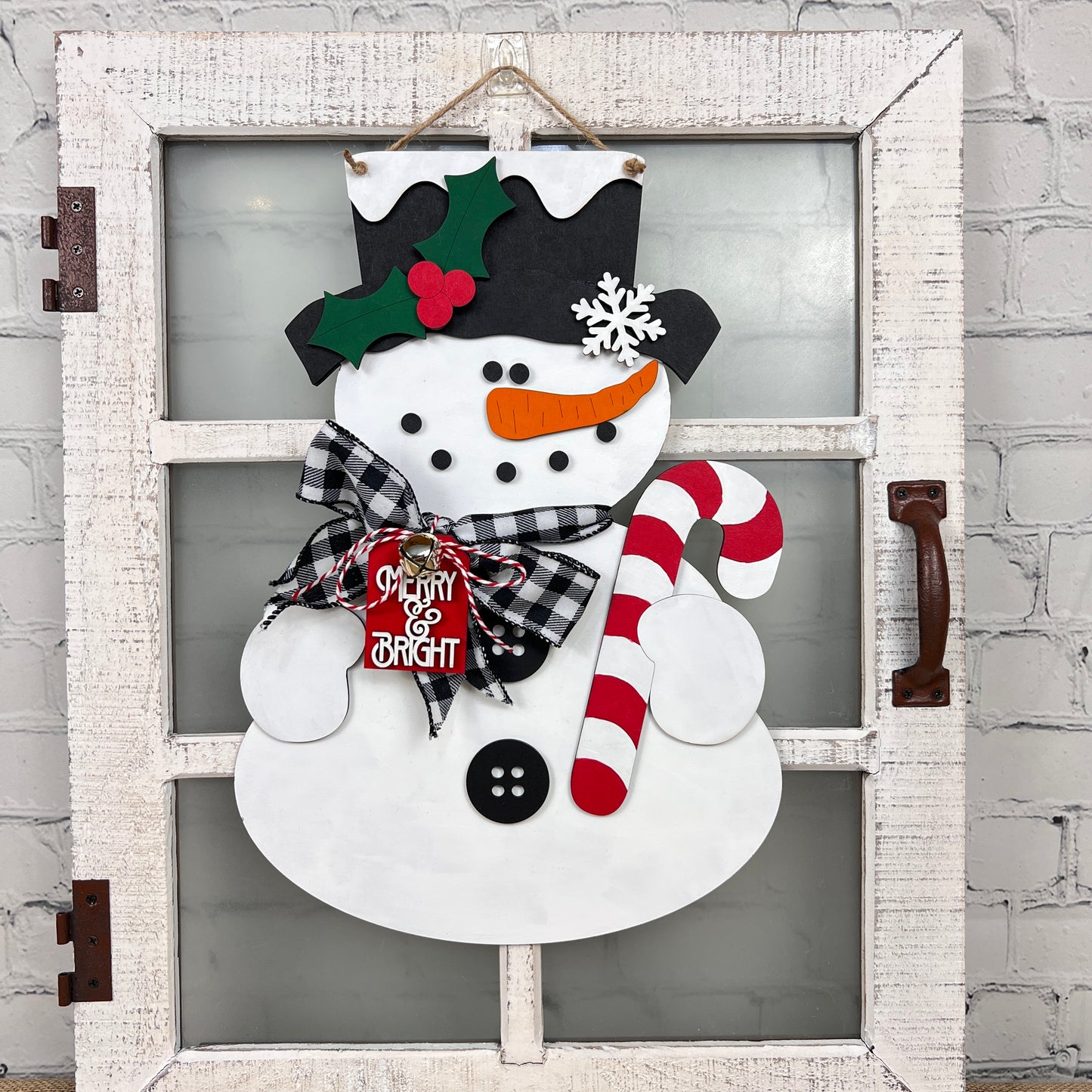 Decorate your home with the Snowman Door Hanger from Janet's Craft Corner, featuring a festive snowman on a rustic window pane backdrop. This charming door hanger showcases a black hat adorned with holly, a plaid scarf, and holds a candy cane. A small "Merry & Bright" sign adds an extra touch of cheer to its chest. It's perfect for enhancing your décor against any brick wall background.