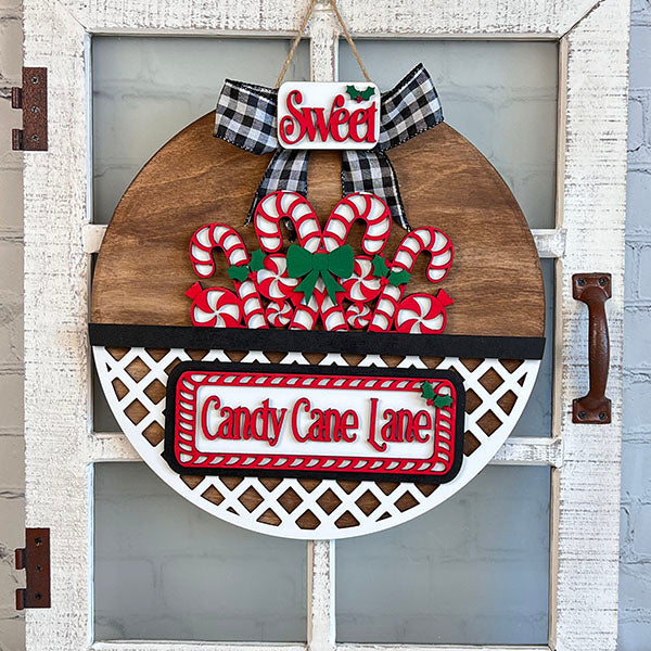 This "Candy Cane Lane" interchangeable insert from Janet's Craft Corner is a vibrant hand-painted element in a DIY home decor kit. It showcases vivid red and white candy cane decorations, complete with a black and white checkered bow. The word "Sweet" is beautifully highlighted at the top, surrounded by holly accents against a rustic wooden door backdrop.