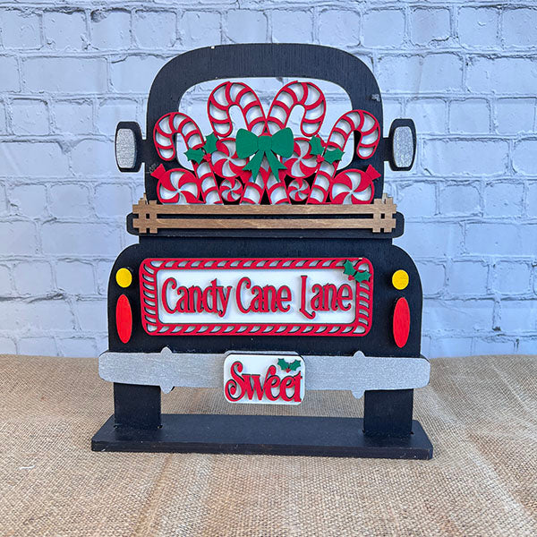 Displayed against a white brick wall is a decorative black truck adorned with candy canes and green bows, showcasing Janet's Craft Corner's hand-painted DIY home decor kit, the "Candy Cane Lane Interchangeable Insert," featuring signs that read "Candy Cane Lane" and "Sweet.