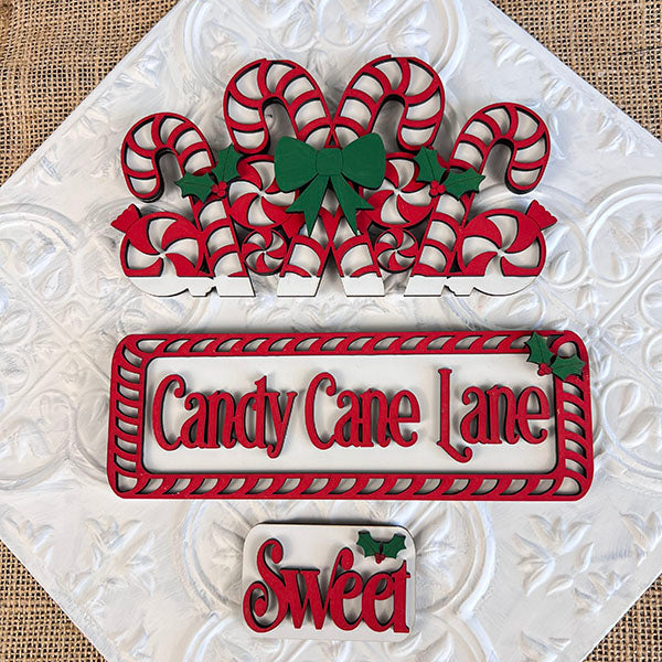 Immerse yourself in the charm of Candy Cane Lane with the festive decorations from Janet's Craft Corner. This DIY home decor kit includes eight candy canes embellished with green bows and holly. A sign displaying "Candy Cane Lane" is framed with red and white stripes, while a smaller "Sweet" sign features holly on a textured background—perfect for your hand-painted projects.