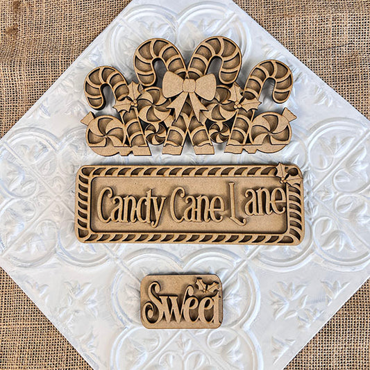 Displayed on a textured background are three wooden craft pieces from Janet's Craft Corner: a hand-painted DIY home decor kit featuring candy canes with a bow, an interchangeable insert titled "Candy Cane Lane," and a smaller piece embellished with the word "Sweet" and decorative leaves.