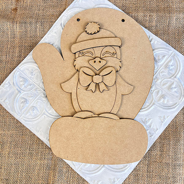 The Penguin Mitten Sign from Janet's Craft Corner showcases a penguin shape with a hat and bow, set on a textured white mat over burlap. It's perfect for DIY kit enthusiasts wanting to personalize their home décor with charming flair.