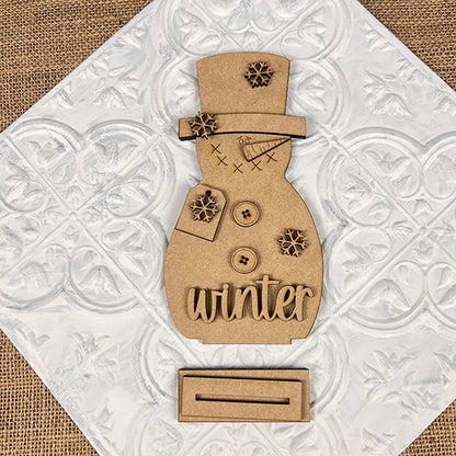 Experience cozy home décor with the Snowman Shelf Sitter from Janet's Craft Corner. This decorative wooden piece features buttons, snowflake details, and the word "winter" at its base. It rests on a patterned white tile placed on a burlap surface, bringing warmth and charm to your space.