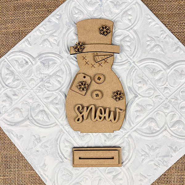 The Snowman Shelf Sitter by Janet's Craft Corner is set against a textured tile background. This charming home décor piece features buttons, snowflakes, and the word "snow," making it perfect for adding a touch of winter magic to any room.