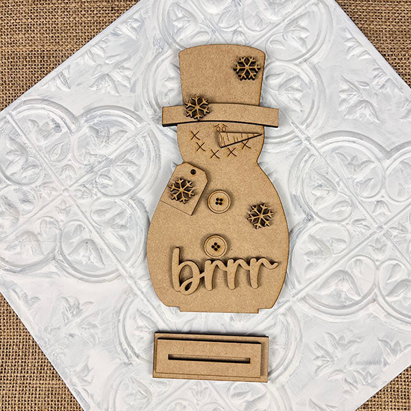 The Snowman Shelf Sitter by Janet's Craft Corner is a delightful DIY kit that includes a wooden snowman proudly sitting on a decorative white tile background. It features charming buttons, snowflakes, and the word "brrr" elegantly written in cursive. Topped off with a top hat and an attached tag, it brings a cozy feel to your home décor.