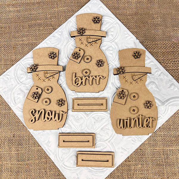 Janet's Craft Corner offers a Snowman Shelf Sitters DIY kit featuring three brown MDF cutouts decorated with snowflakes, buttons, and winter-themed words. These charming seasonal décor pieces rest on a textured white and burlap background for cozy home styling.