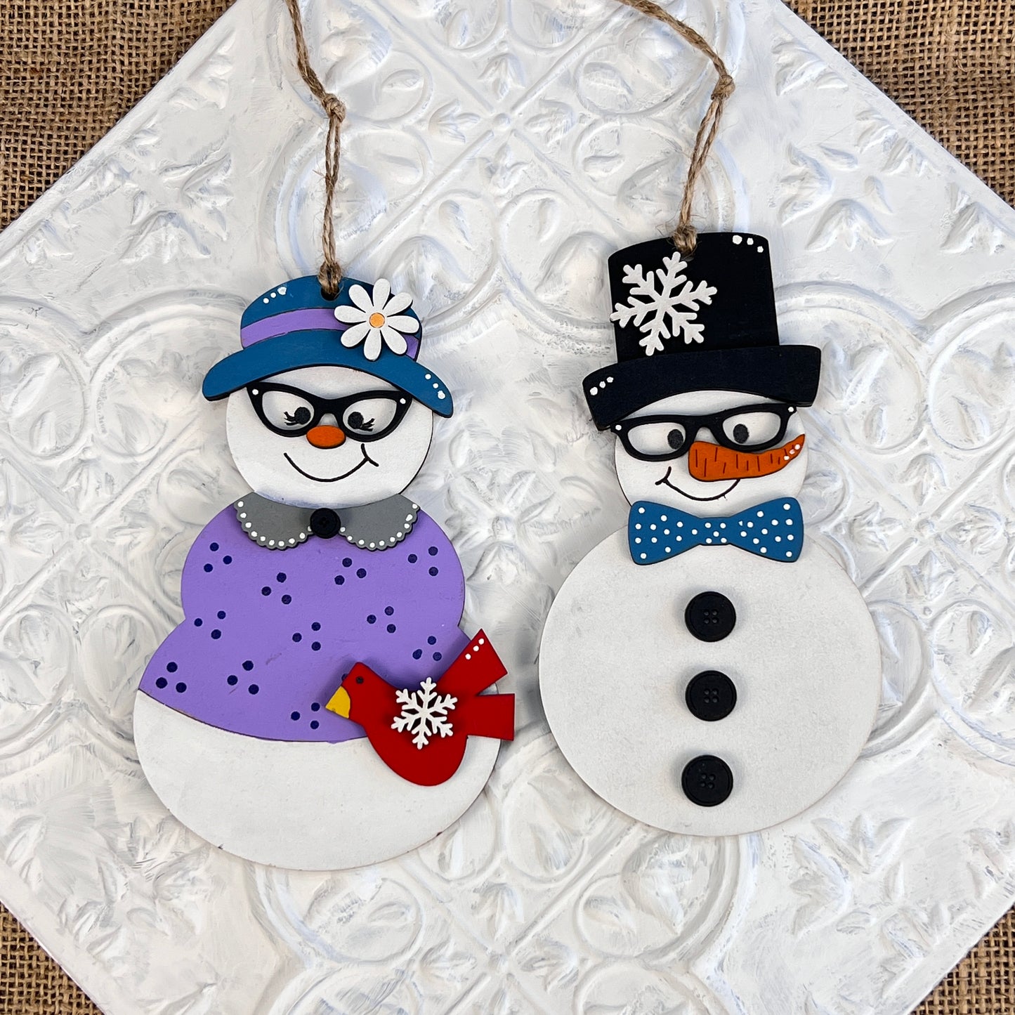 The Vintage Snow Couple Ornaments - Painted by Janet's Craft Corner feature two charming snowman figures on a white textured background. One wears a blue hat and purple scarf, while the other sports a black hat and blue bow tie. Both snowmen have carrot noses, black eyes, and details of snowflakes and buttons that capture the essence of vintage holiday decor.