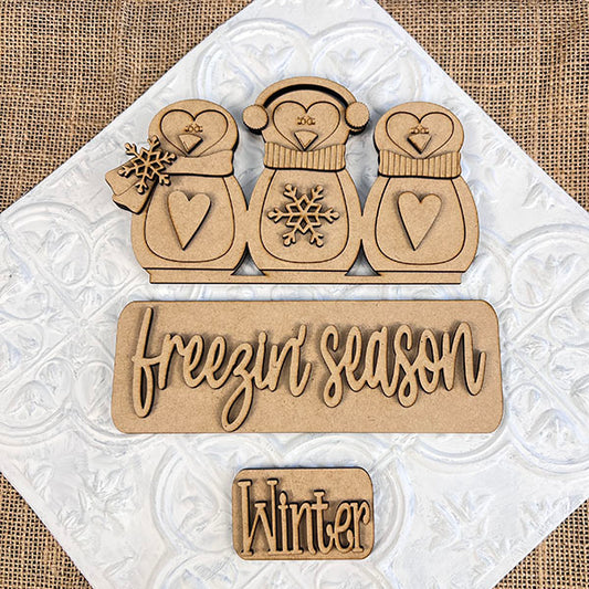 Janet's Craft Corner presents the "Freezin' Season" Interchangeable Insert, a DIY home decor kit featuring wooden cutouts on a textured background with charming penguins wearing winter accessories and decorated with heart-shaped designs. The script "Freezin Season" appears alongside a plaque labeled "Winter," making it an ideal craft kit for versatile home decoration.