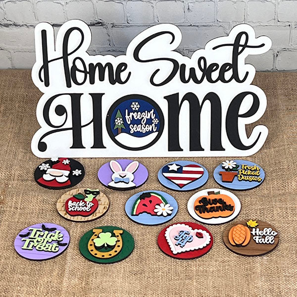 The "Home Sweet Home Interchangeable Set" by Janet's Craft Corner includes a decorative sign with various themed shelf sitters like a snowman, Santa hat, Easter bunny, and more for seasonal decoration.