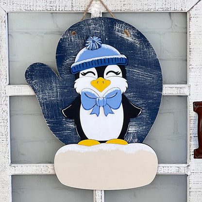 The Penguin Mitten Sign from Janet's Craft Corner is an adorable wooden decoration showcasing a cheerful penguin in a blue hat and bow, joyfully positioned on snow. This delightful piece is mounted against an oval-shaped, dark blue backdrop, making it charming home décor when displayed on a rustic white door.