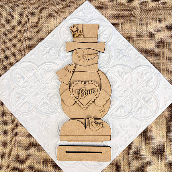 The Valentine Snowman Shelf Sitter by Janet's Craft Corner is a DIY decor craft kit that includes a wooden snowman with heart-shaped "Love" and "Be Mine" details, on a rectangular base with a small bird figure against a textured white and burlap background. Perfect for Valentine's Day decorating.