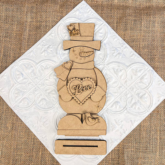 The Valentine Snowman Shelf Sitter by Janet's Craft Corner is a DIY decor craft kit that includes a wooden snowman with heart-shaped "Love" and "Be Mine" details, on a rectangular base with a small bird figure against a textured white and burlap background. Perfect for Valentine's Day decorating.