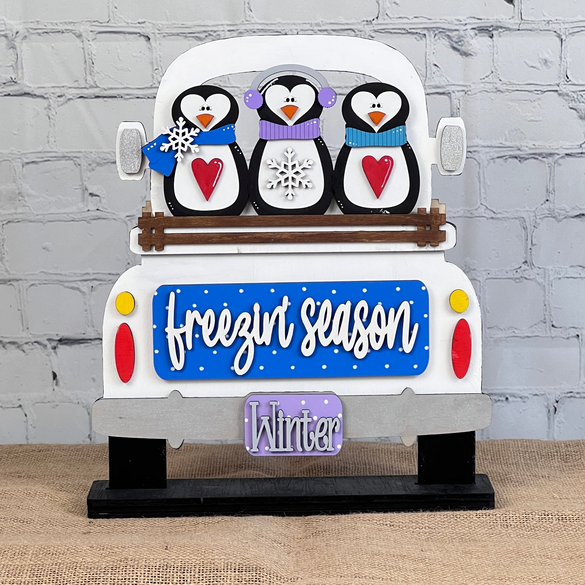 The Freezin' Season Interchangeable Insert from Janet's Craft Corner features playful penguins with heart-shaped bellies sitting in a white truck. The design includes the phrase "freezin' season" on a blue background and a purple "Winter" license plate. This DIY home decor kit creates the perfect accent against a light brick wall.