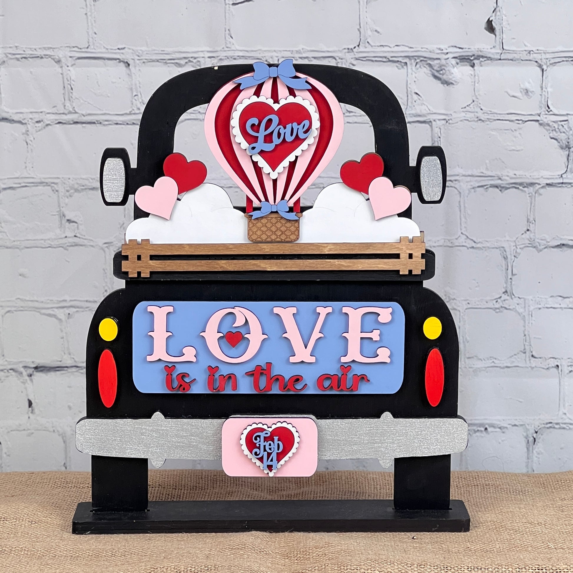 This hand-painted DIY home decor kit from Janet's Craft Corner features the "Love is in the Air" interchangeable insert, showcasing a decorative car-shaped sign themed around love. It includes a hot air balloon adorned with hearts and displays "Feb 14" on the license plate, all set against a backdrop of a white brick wall. It's perfect for celebrating love throughout the year as versatile home decor.