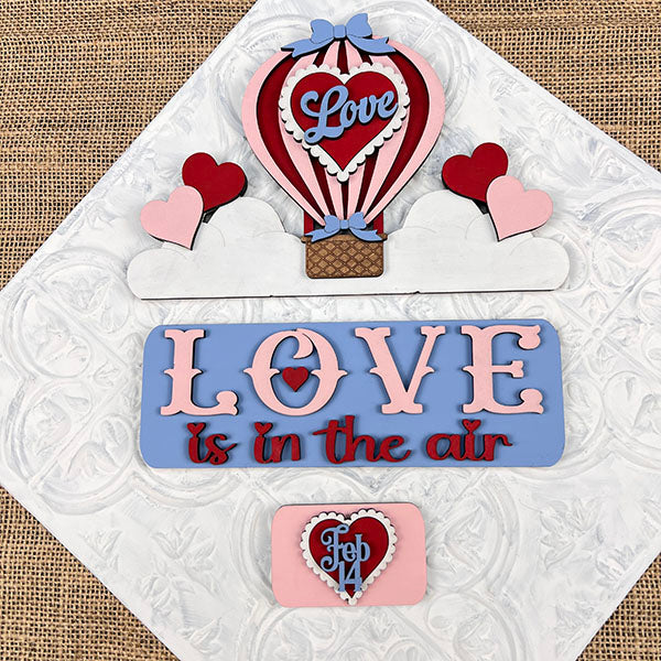 Experience the charm of Janet's Craft Corner with the "Love is in the Air Interchangeable Insert." This DIY home decor kit features decorative cake toppers on a textured surface, showcasing a hot air balloon labeled "Love," hearts, and pastel-colored phrases like "Love is in the Air" and "Feb 14" for an irresistible decorative accent.