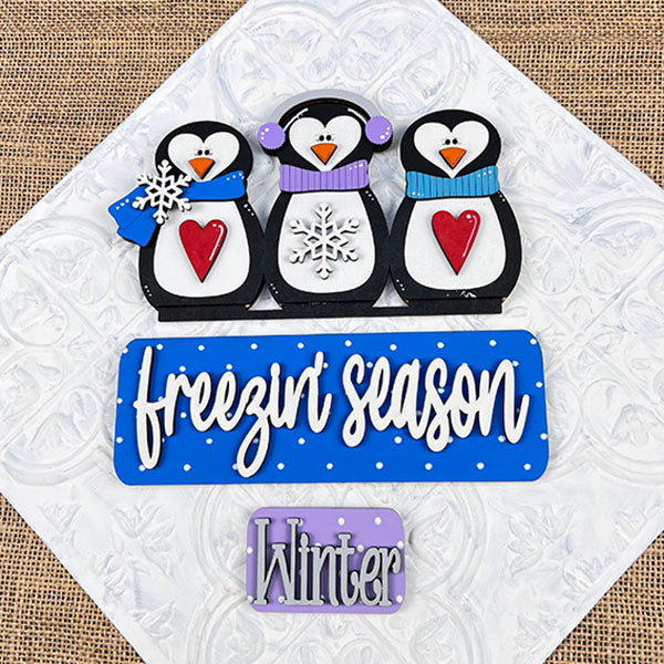 The Freezin' Season Interchangeable Insert by Janet's Craft Corner features three playful penguin figures dressed in charming hats and scarves, each holding colorful hearts and snowflakes. The decorative square showcases a blue sign with the text "freezin' season" above another purple sign labeled "Winter." This delightful DIY home decor kit is perfect for adding versatility to your home styling.