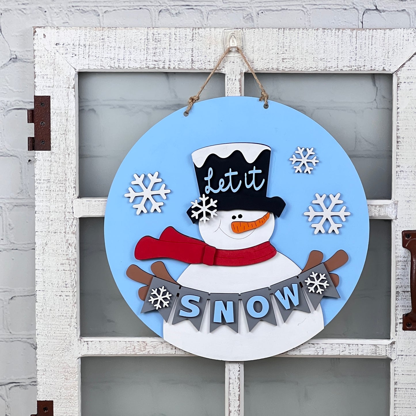 Add a touch of holiday charm to your rustic white door with Janet's Craft Corner's Snowman Let It Snow Door Hanger. This delightful round blue decoration showcases a snowman wearing a top hat and red scarf. The words "Let it" embellish the hat, while "SNOW," surrounded by whimsical snowflakes, is prominently displayed below.