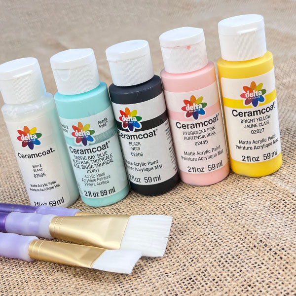 Five bottles of Janet's Craft Corner paint in white, green, black, pink, and yellow are lined up on a burlap surface. Perfect for your DIY kit projects, two paintbrushes with purple and white handles are laid in front of the bottles alongside the Spindle Birdhouses Shelf Sitter.