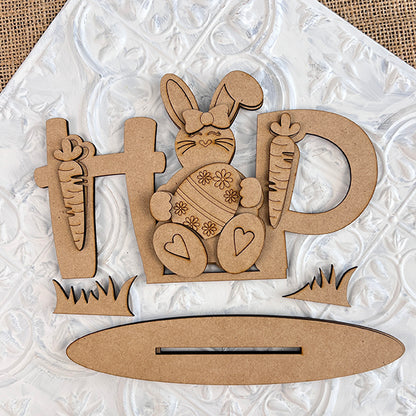 The Easter Trio Mini Shelf Sitters by Janet's Craft Corner is a delightful DIY kit that includes a wooden cutout spelling "HOP," featuring a bunny sitting on the "O." This set is perfect for Easter decor, embellished with floral patterns and heart shapes, with the bunny holding a carrot. Additional carrots and grass elements are included, all set against a textured white background for charming mini shelf sitters.