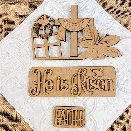 Janet's Craft Corner offers the "He is Risen Interchangeable Insert" DIY home decor kit, which showcases wooden cutouts of a draped cross, a butterfly, and a window featuring a dove and wreath. The kit includes two plaques with the messages "He is Risen" and "Faith," designed to be interchangeable on a textured white and burlap background for an Easter-themed craft experience.