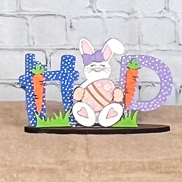 Create your own festive scene with the Easter Trio Mini Shelf Sitters from Janet's Craft Corner. This delightful DIY kit includes charming decor featuring the word "HOP," where a sitting cartoon bunny and carrots cleverly form the letter "O." The textured blue and purple letters are designed to rest on a green grass base, making it an ideal addition to your shelf decor.