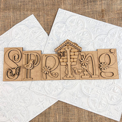 The "Spring Word Block Shelf Sitter" from Janet's Craft Corner is a DIY seasonal home decor kit featuring wooden blocks with floral and nature motifs, including butterflies and birds. Ideal for home decor, these blocks look stunning on textured white tiles atop a burlap surface.