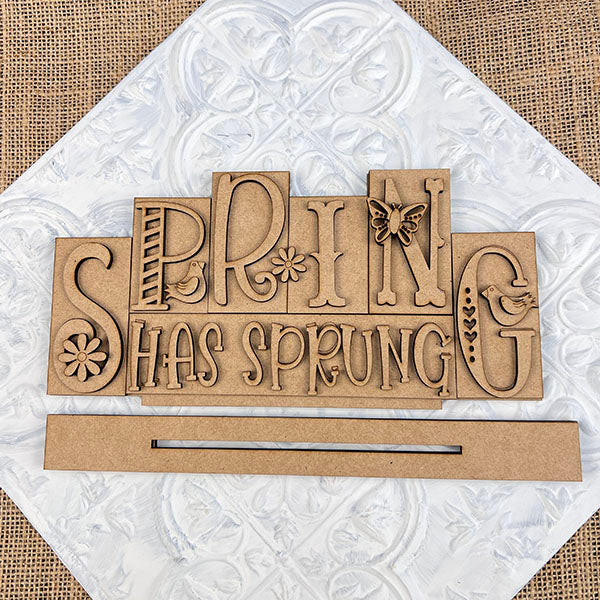 The "Spring Has Sprung" Mini Word Block Shelf Sitter by Janet's Craft Corner is a decorative wooden piece featuring floral and butterfly designs on a textured white and burlap background, perfect for seasonal home decor.