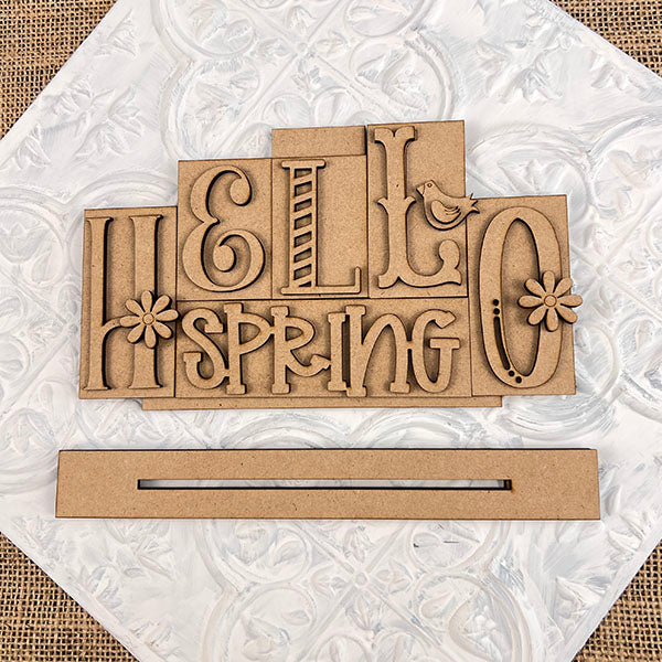 The Hello Spring Mini Word Block by Janet's Craft Corner is a DIY seasonal home decor craft kit. Featuring the words "HELLO SPRING" with decorative lettering, flowers, and a bird above a rectangular base, it's perfect against textured white backgrounds for creative projects. Set of 1 included.