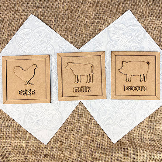 The Bacon Eggs Milk Signs DIY kit from Janet's Craft Corner includes three wooden signs on a burlap and patterned white background, with rustic animal engravings: a chicken for "eggs," a cow for "milk," and a pig for "bacon"—ideal for farmhouse-style décor lovers.