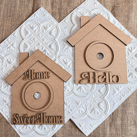 Janet's Craft Corner offers a "House Interchangeable Set," a DIY home décor kit with decorative wooden house-shaped plaques. One says "Home Sweet Home," the other "Hello," both have center cutouts. The set includes 1 base and 12 inserts, ideal for craft enthusiasts seeking personalized decor.