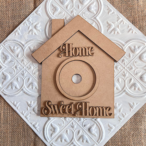 This delightful wooden decoration, from the House Interchangeable Set by Janet's Craft Corner, is house-shaped and sits atop a decorative white tile background. It features the words "Home Sweet Home" in raised letters along with a small circular opening at its center, enhanced by the textured background that complements its home décor charm.