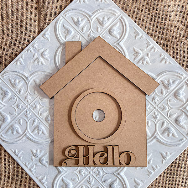 The House Interchangeable Set by Janet's Craft Corner features a delightful wooden birdhouse cutout with "Hello" inscribed at the bottom, beautifully displayed on a textured white tile adorned with an intricate leaf pattern. Set against a rustic burlap backdrop, this piece is part of a hand-painted DIY craft kit that makes for an ideal addition to your home décor collection.