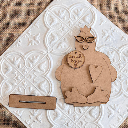 The Farm Standing Shelf Sitter from Janet's Craft Corner is a charming wooden cutout shaped like a cartoon chicken wearing glasses, adorned with heart motifs and featuring a sign that reads "Fresh Eggs 5¢." It provides a delightful rustic touch and is showcased on a white textured background alongside a rectangular piece with a slot, making it ideal for farmhouse decor enthusiasts.