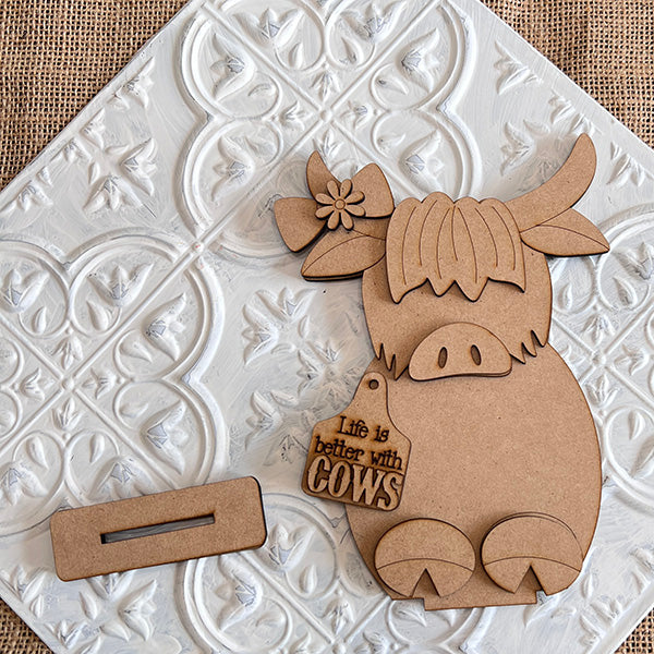 The "Farm Standing Shelf Sitter" by Janet's Craft Corner features a wooden cutout of a cow adorned with a flower and a tag that says "Life is better with cows," providing a charming rustic touch to your decor. Ideal for farmhouse-inspired spaces, it rests on a textured white tile surface, complemented by an adjacent rectangular wooden piece.