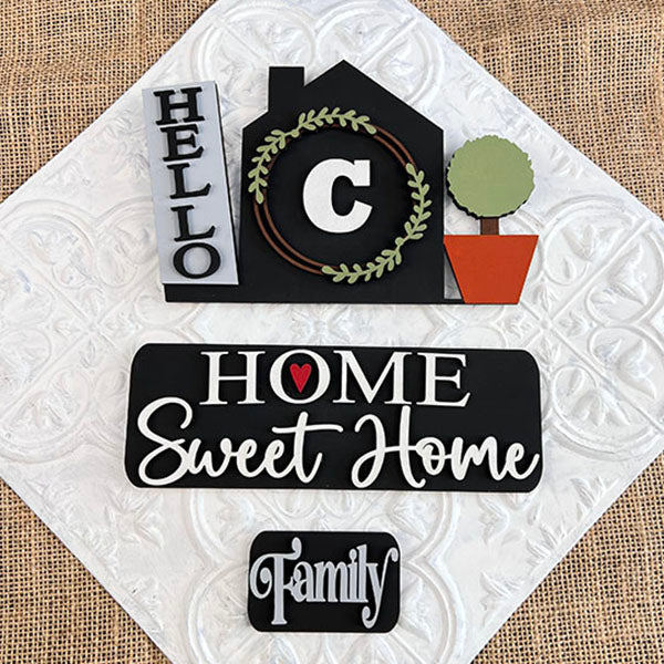 The "Hello Home Monogram Interchangeable Insert" from Janet's Craft Corner is a decorative sign featuring the phrases "Hello," "Home Sweet Home," and "Family." It is designed in the shape of a house with a letter "C," complemented by a green wreath and a small potted plant on a textured white background, making it an ideal piece for versatile home decor.